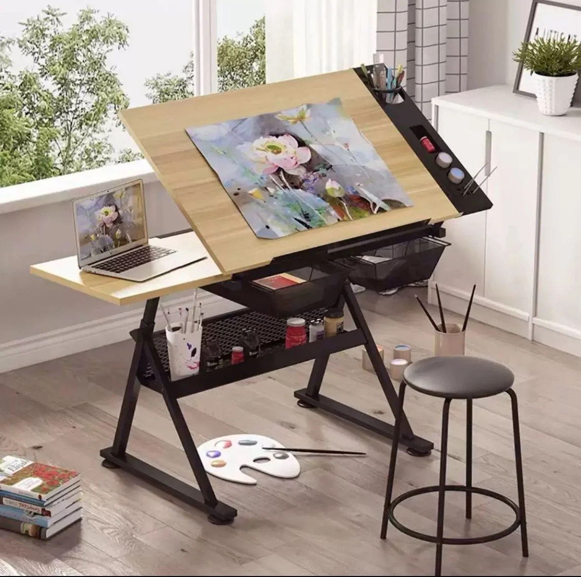 طاولة رسم احترافية سوداء مع سطح خشبي مائل يضم حاسوب محمول وأدوات رسم وكرسي دائري صغير بجانبها
