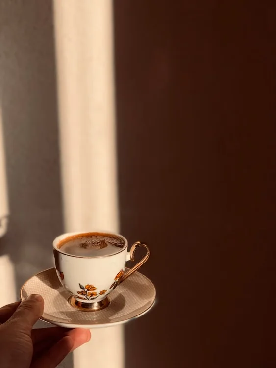 فنجان قهوة أبيض مزين بنقوش زهور صغيرة مع حافة ذهبية وصحن متناسق محمول باليد تحت ضوء الشمس.