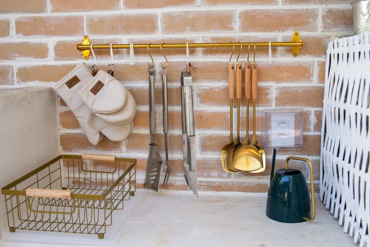 سكة معدنية ذهبية معلقة على جدار قرميدي، تحتوي على أدوات مطبخ متنوعة وقفازات فرن بيضاء وسلة تخزين جانبية أنيقة.